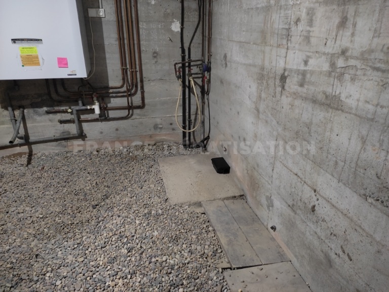 Mur béton, chauffe-eau et tuyaux, sol gravillonné et poste noir